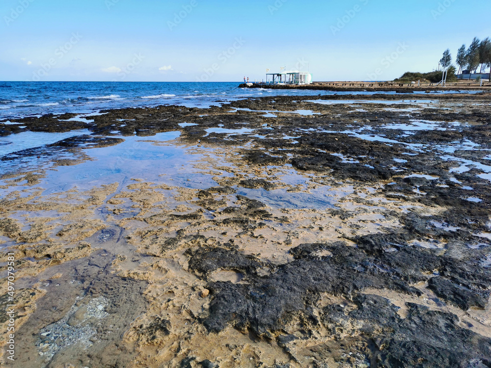 Fototapeta premium The rocky coast of the Mediterranean Sea from long-hardened lava, waves, clear water against a blue sky with clouds.