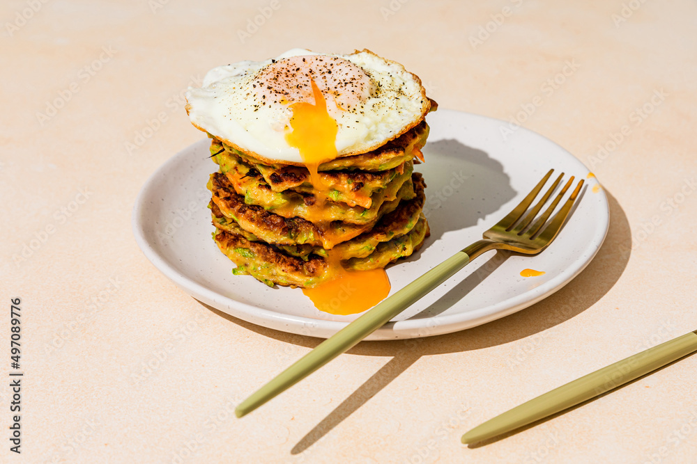 Close-up of a stack of fried vegetable pancakes, made from zucchini and carrot, with a fried egg with runny yolk on top, shot with hard light with shadows, served on a plate with golden knife and fork - obrazy, fototapety, plakaty 