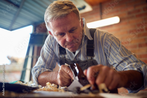 Mature Male Carpenter In Garage Workshop Planing Piece Of Wood