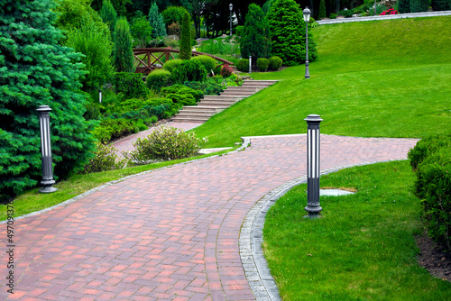 iron ground lantern and poli garden lighting of park curved path paved with stone tiles with drain lattice in park among plants, evergreen bushes and pine trees on slope hill green lawn, nobody. photo