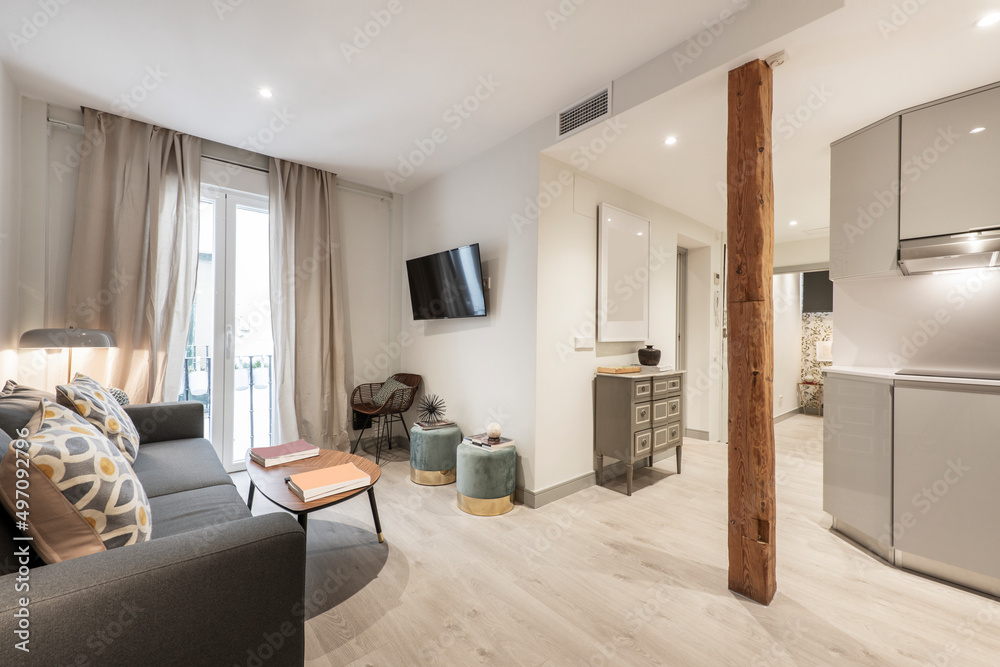 Living room with three-seater sofa, wooden coffee table, balcony with curtains and net curtains, wooden column and kitchen with gray furniture Living room with three-seater sofa, wooden coffee table, balcony with curtains and net curtains, wooden column and kitchen with gray furniture