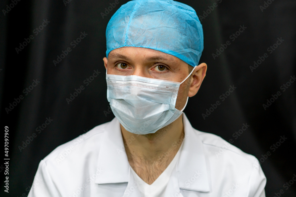 Portrait of a young doctor on a black background. A surgeon in a white coat, wearing a medical mask and a surgical cap, looks carefully to the side.