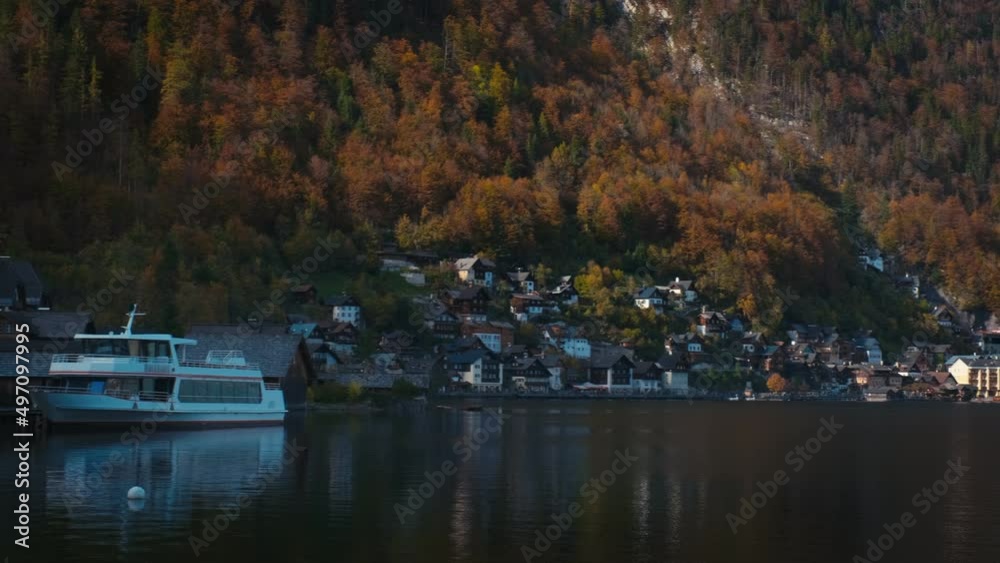 Autumn fall in famous tourist destination serene town Hallstatt in mountains Alps. Yacht, Cathedral church, houses, golden leaves on trees reflected in Hallsttattersee. Austria. Horizontal camera pan