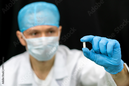 A doctor in a white coat holds a pill in his hand on a black background. Medical preparations for the treatment of coronavirus. A young intern doctor is considering a new drug.