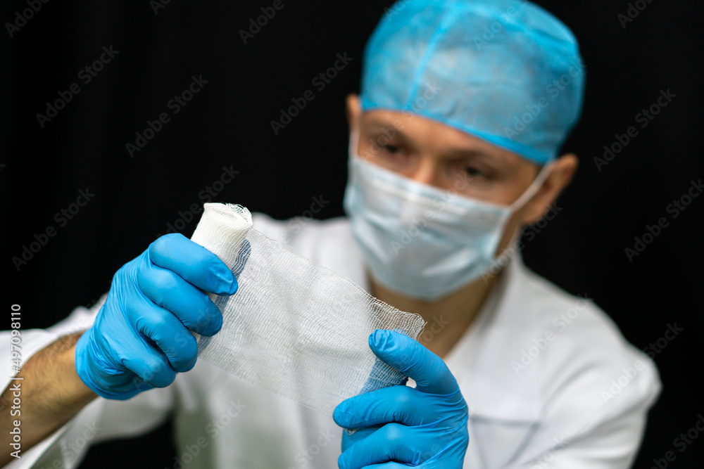 A young traumatologist holds a white medical bandage in his hand. A doctor in a white coat and a bandage in his hands on a black background. A young traumatologist holds a white medical bandage in his hand. A doctor in a white coat and a bandage in his hands on a black background.