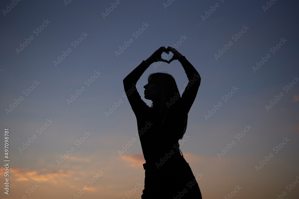 Attractive caucasian long hair female posing in front of the camera in silhouette showing love or heart gesture outdoor at sunset with sky view. Romantic or love concept. High quality image Attractive caucasian long hair female posing in front of the camera in silhouette showing love or heart gesture outdoor at sunset with sky view. Romantic or love concept. High quality image