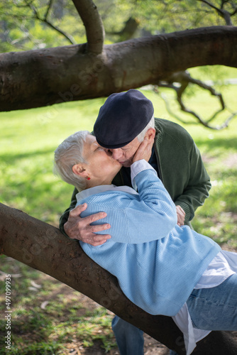 Romantic elderly husband and wife kissing in park. Happy man and woman hugging kissing when woman lying on thick tree branch and enjoying rest. Love, relations and active rest of aged people concept
