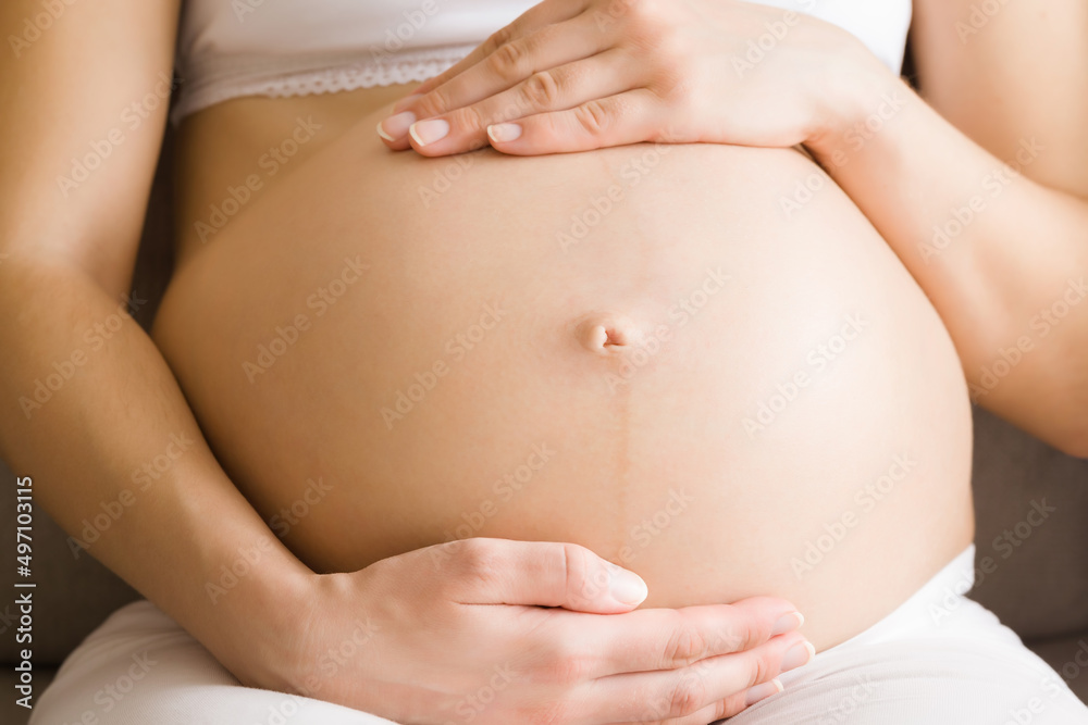 Young adult pregnant woman sitting and touching naked big belly and feeling her baby move. Emotional loving pregnancy time. Baby expectation. Front view. Closeup.