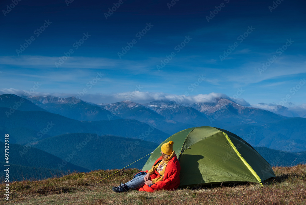 A young girl sitting near the camp tent with mountains peaks and blue sky on background. Happy girl resting in campsite