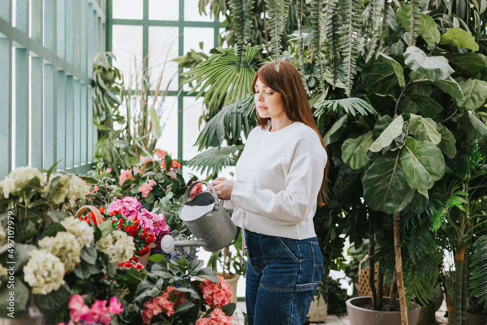 young woman plus size model waters flowers from watering can in greenhouse with tropical plants and potted plants, spring concept of urban and home jungles, place for rest and relaxation mental health