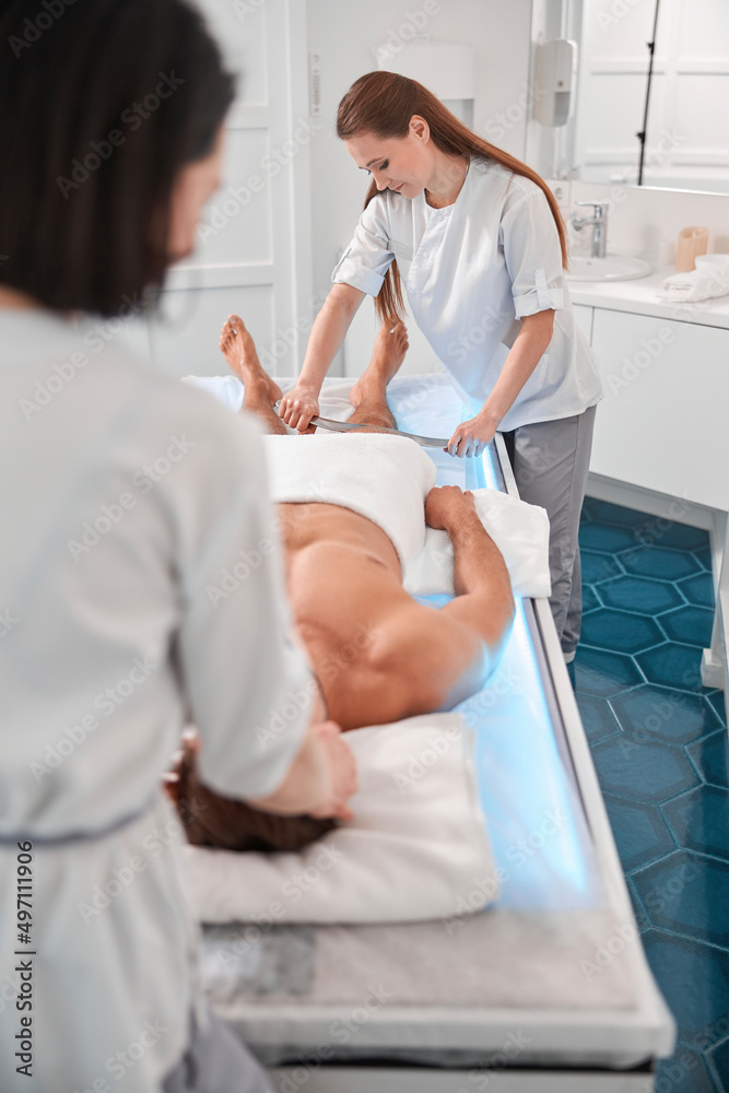 Therapist uses tool to massage man client leg working with skilled colleague in hospital office Therapist uses tool to massage man client leg working with skilled colleague in hospital office