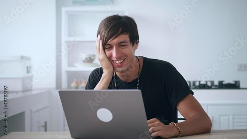 Young happy man looking at laptop monitor and laughing while reading funny news while sitting at kitchen table at home. Smiling male watches funny videos on the Internet and laughs at what he saw