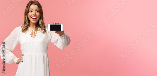 Portrait of cheerful, upbeat attractive blond caucasian girl in white dress, showing smartphone display, hold mobile phone horizontal smiling camera amazed, stand pink background