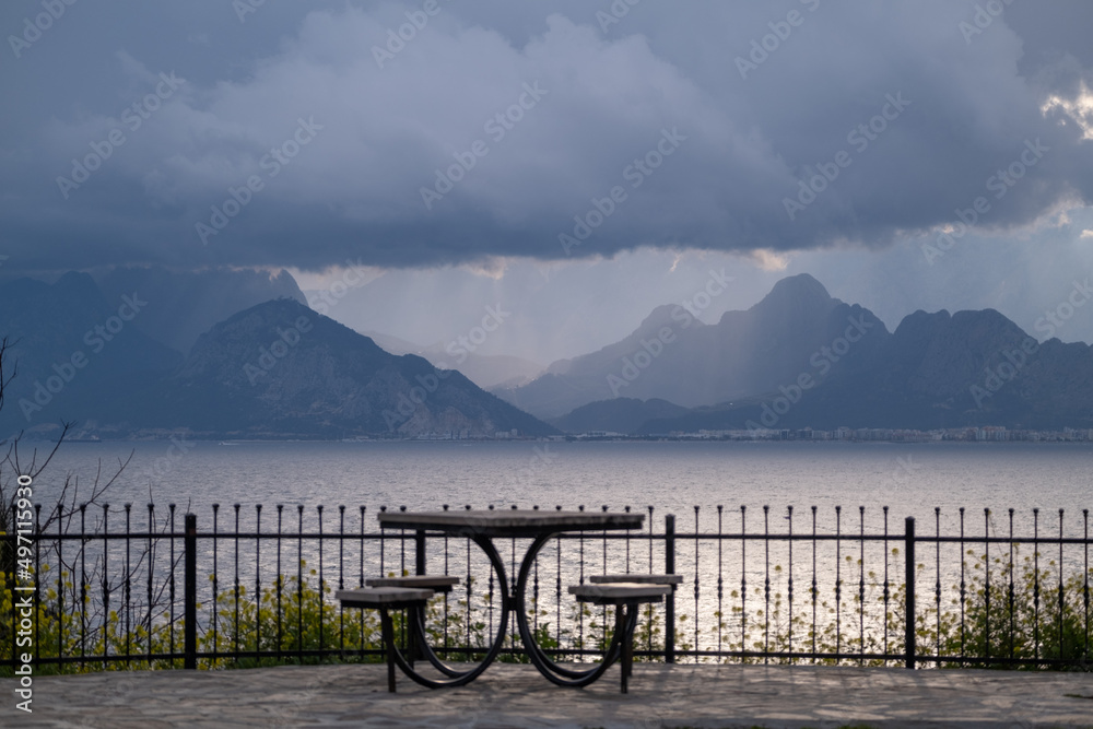 A table and chairs as a place to relax in a city park against the mountains and a hazy, sea and an incredible sky with clouds A table and chairs as a place to relax in a city park against the mountains and a hazy, sea and an incredible sky with clouds