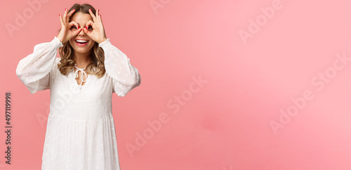 Portrait of dreamy cute and funny young blond girl seeing something interesting, looking from okay signs as making glass-mask with fingers over eyes, smiling amused, pink background