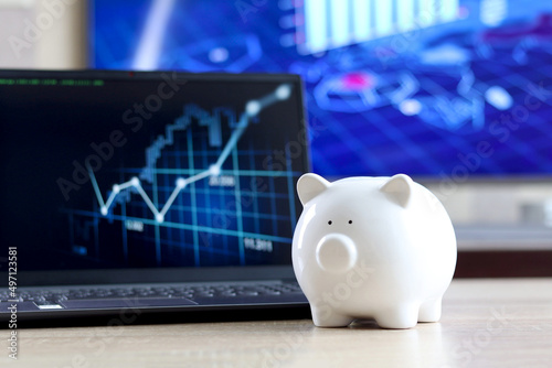 A white piggy bank on a laptop displaying an online financial chart and graph used as an online stock trading, asset allocation or financial analysis and e-commerce concept.