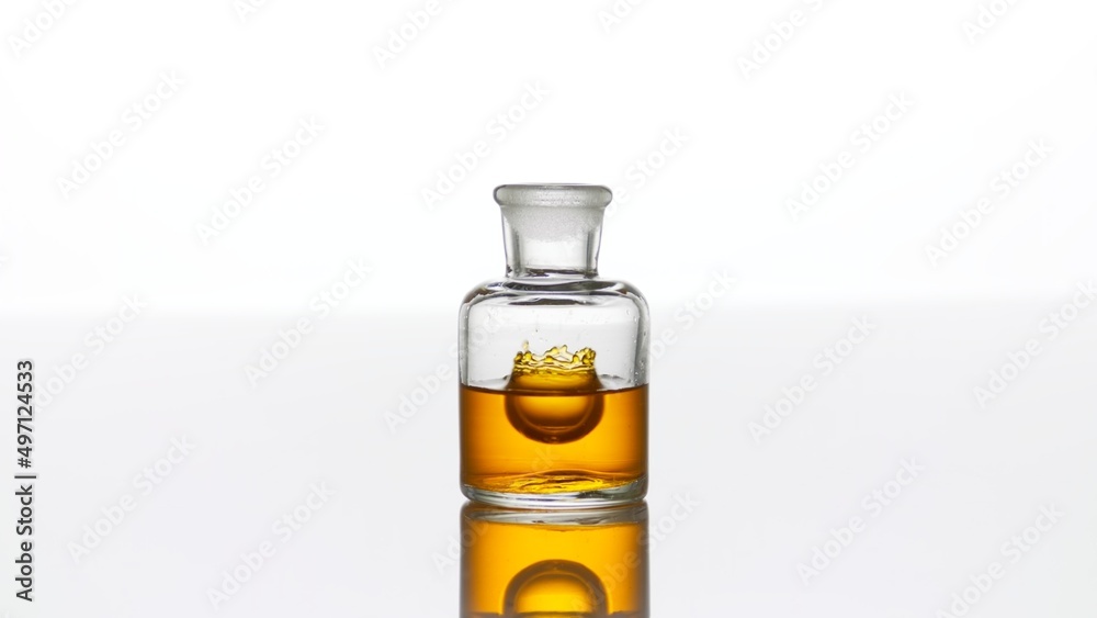 Drop of argan oil falls down into clear old fashioned bottle with yellow transparent liquid on white background making crown inside it | Abstract cosmetics formulation concept Drop of argan oil falls down into clear old fashioned bottle with yellow transparent liquid on white background making crown inside it | Abstract cosmetics formulation concept