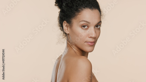 Young slim gorgeous African American woman with bun hairstyle turns and looks at camera over her bare shoulder on beige background | Beauty care concept