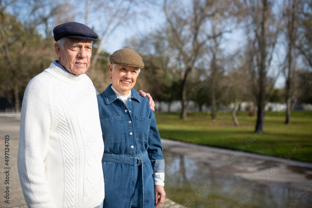 Portrait of happy aged couple spending day in park. Smiling man and woman resting in city recreation zone standing close hugging and looking aside. Elderly couples relations and active rest concept Portrait of happy aged couple spending day in park. Smiling man and woman resting in city recreation zone standing close hugging and looking aside. Elderly couples relations and active rest concept