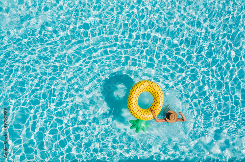 Top view of a young female in swimsuit bikini in a straw hat on blue swimming pool waves background with big inflatable Yellow Pineapple tube. Chill out a summer vacation in luxury resorts concept. #497131989