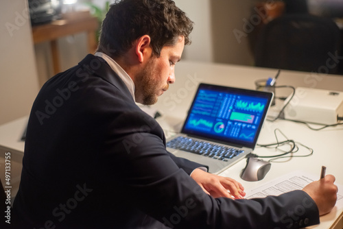 Back view of thoughtful young man working. Man in suit sitting at laptop with charts on screen, writing, working late. Business, job, occupation, late night work concept