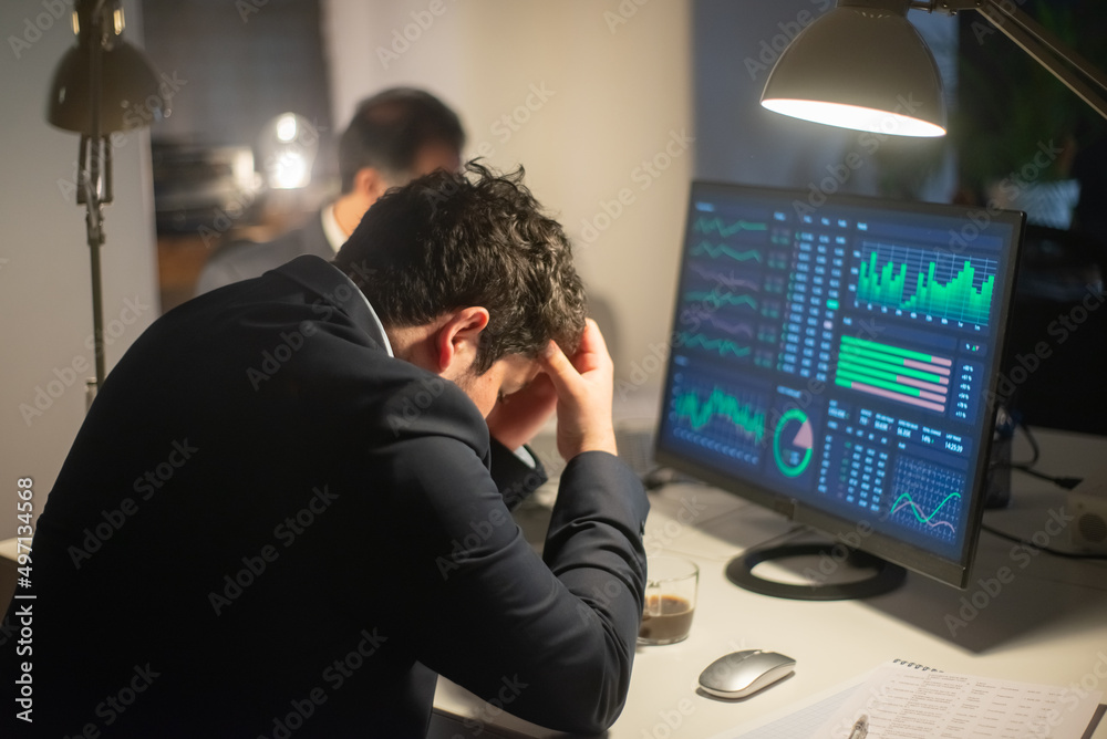 Back view of upset young man working. Man in suit sitting at computer with charts on screen, holding head in distress. Business, job, occupation, late night work concept Back view of upset young man working. Man in suit sitting at computer with charts on screen, holding head in distress. Business, job, occupation, late night work concept
