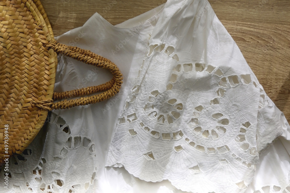 Fototapeta premium White linen lace blouse and round straw bag on wooden background. Flat lay.