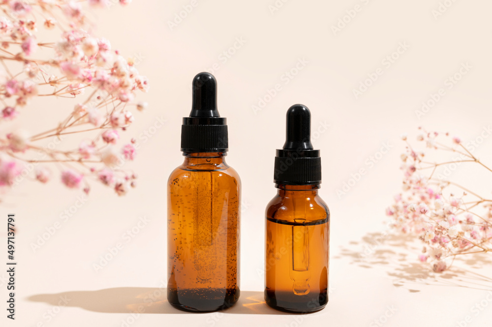Brown glass cosmetic bottles with pipette, dry pink flowers on beige background with sun shadows. Natural organic cosmetics for face and body, aroma oil. Beauty concept. Products for branding, mock up