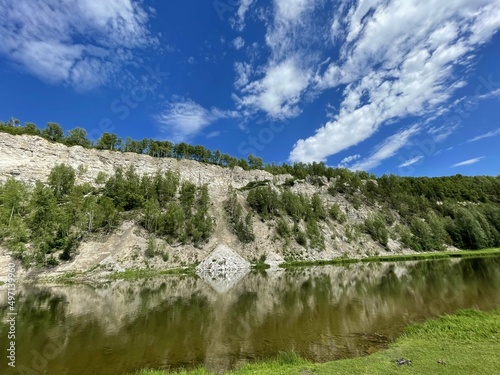 View of the other side of the river, on the other side of the white rocks with sprouted bushes of plants. The rocky shore is reflected in the river. A sunny summer day with a soft blue sky and clouds.