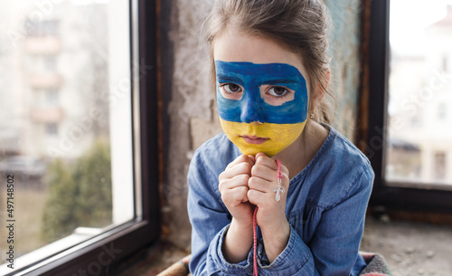 a little Ukrainian patriot girl with the flag of Ukraine on her face prays sitting at the window. Children are against the war in Ukraine photo
