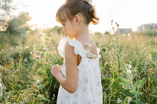 Adorable little girl wearing natural white dress with wildflower motiv in green field with wild carrot flowers at summer  outdoor lifestyle. Freedom concept