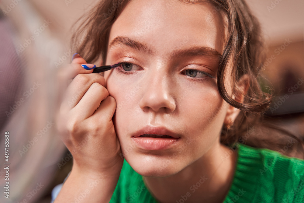 Teenage girl holding brush with eyelids and looking attentively at the mirror