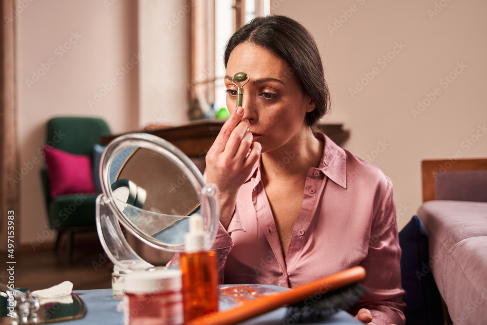 Female is sitting in front of mirror in living room and using jade roller on her face Female is sitting in front of mirror in living room and using jade roller on her face