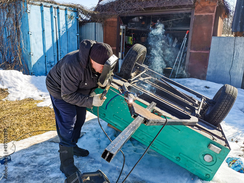 An elderly male welder repairs a car trailer with electric arc welding on a spring sunny day.