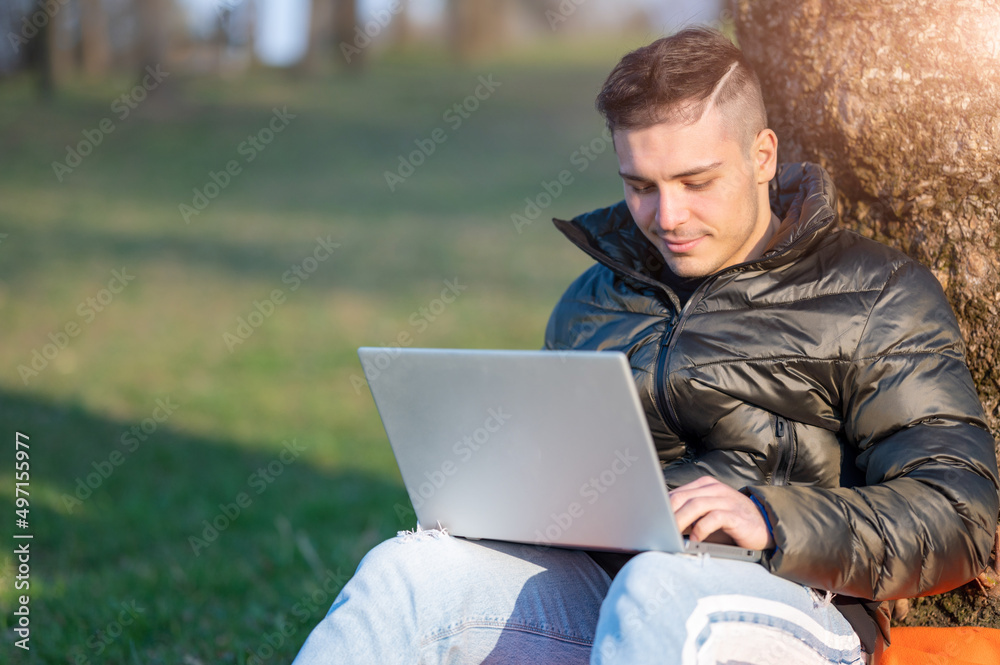 Focused handsome man in casual wear working with laptop outdoors. Sitting at the foot of a tree, he enjoys nature on a sunny spring day. Remote working concept. Copy space.