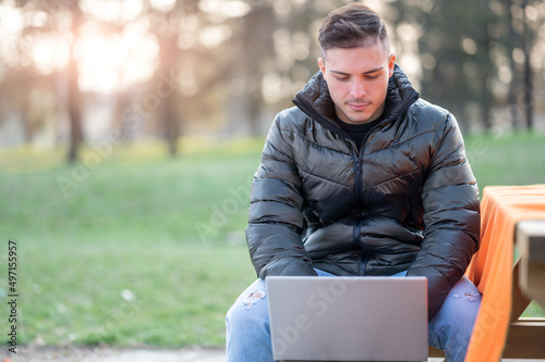 A attractive young office worker in casual wear, is focused.while typing on his laptop. I work remotely on a bench in the park. Remote working outdoors concept.