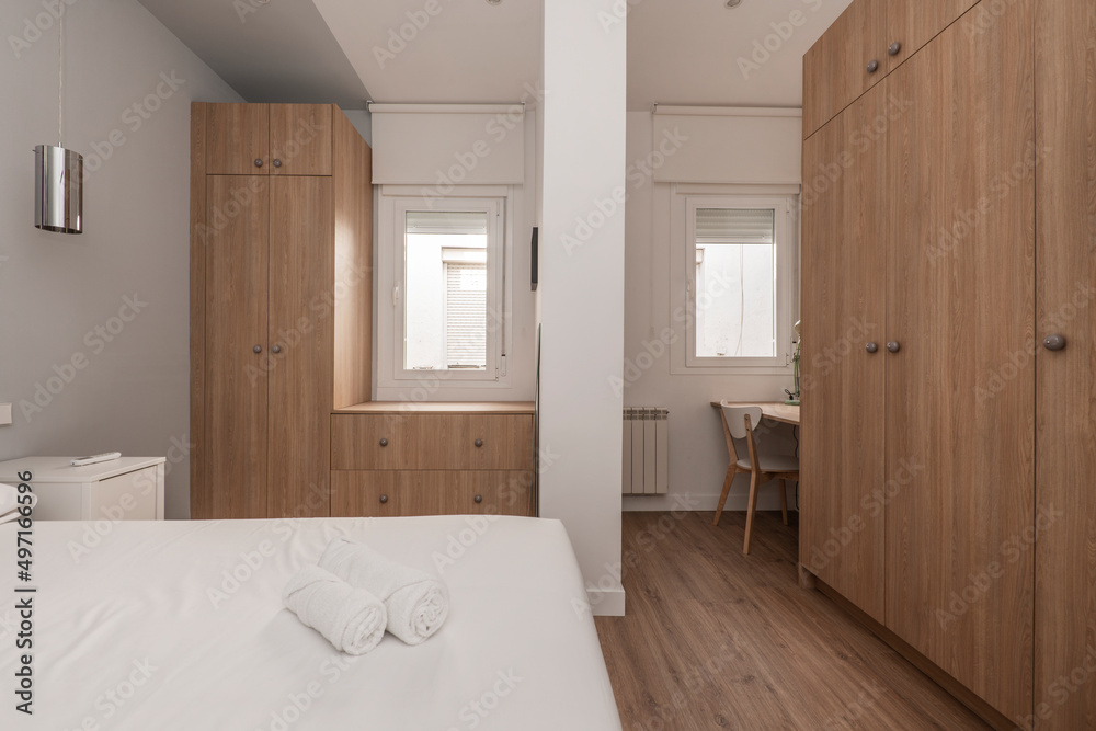 Bedroom with double bed, with white bedding, cabinets with doors in wood tones, desk with chair, chest of drawers, white bedside tables and several white aluminum windows Bedroom with double bed, with white bedding, cabinets with doors in wood tones, desk with chair, chest of drawers, white bedside tables and several white aluminum windows