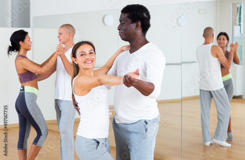 Young smiling Latina enjoying slow foxtrot with african american partner in dance studio for adults