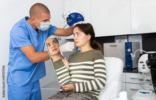 Female client looking at mirror while qualified beautician examining her face skin after procedure in medical cosmetology office..