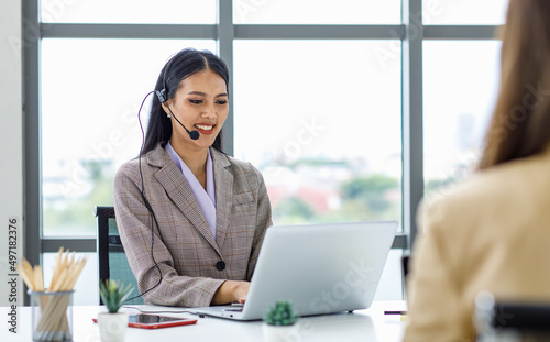 Asian young beautiful happy female businesswoman customer service call center operater in formal business suit sitting at working desk chat online with client via microphone headset in company office