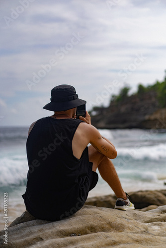 A man traveler on a rock by the ocean uses a phone.