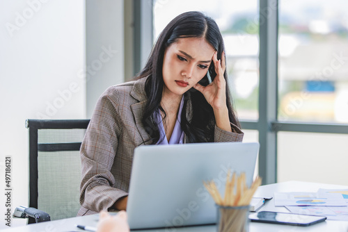 Asian young unhappy upset stressed depressed frowning face female businesswoman employee staff in formal business suit sitting at working desk holding hands on head having problem trouble headache