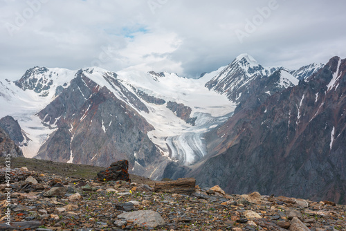Dramatic landscape with high snowy mountain range with long glacier tongue and pointed peak in sunlight under cloudy sky. Atmospheric view from stone hill to large snow mountains at changeable weather