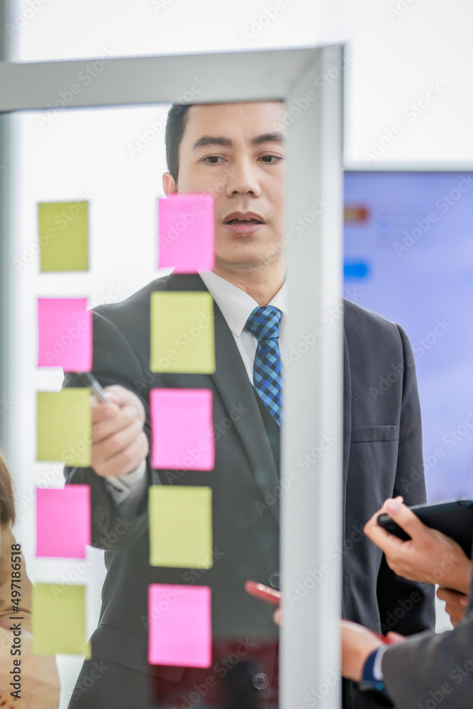 Asian happy male and female professional successful businessman and businesswoman colleagues partnership teamwork in formal business suit standing smiling look at post it note on glass board together Asian happy male and female professional successful businessman and businesswoman colleagues partnership teamwork in formal business suit standing smiling look at post it note on glass board together