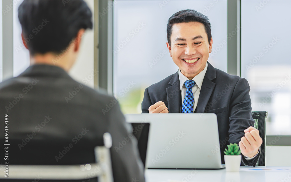 Closeup shot Asian happy professional successful male businessman employee in formal business suit sitting smiling holding fist up celebrating customer job goal agreement done achievement in office Closeup shot Asian happy professional successful male businessman employee in formal business suit sitting smiling holding fist up celebrating customer job goal agreement done achievement in office