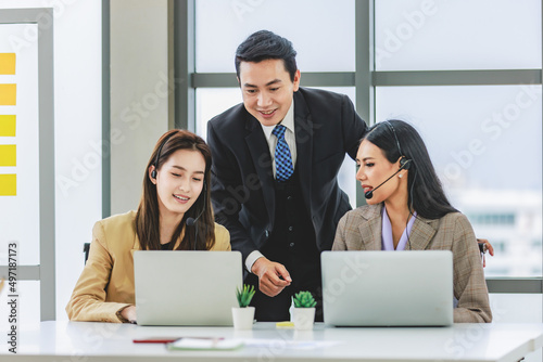 Asian male businessman manager teaching two millennial young beautiful female customer service call center in formal business suit wearing microphone headset sitting working talking with customers