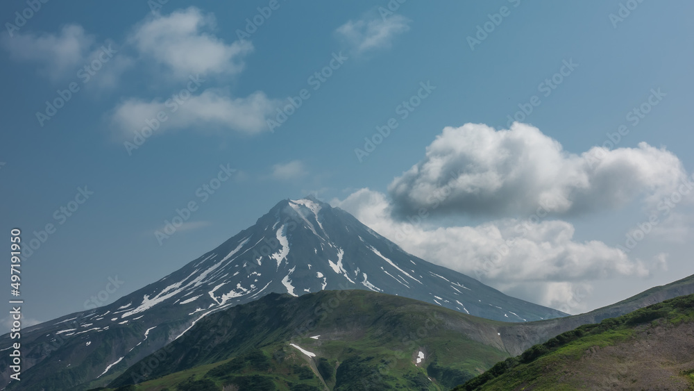 A beautiful conical volcano on a blue sky background. Snow on the slopes. Clouds at the top. In the foreground are green hills. Kamchatka. Stratovolcano Vilyuchinsky
