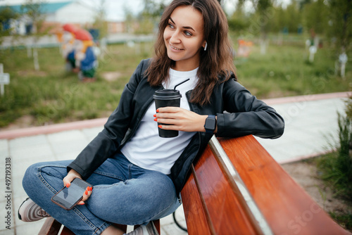 beautiful brunette in a leather jacket sits on a bench in the park and drinks takeaway coffee. enjoying the outdoors
