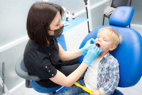 A cute little boy sits at the dentist, opened his mouth, showing his teeth to the dentist. Happy boy is sitting in the dentist's chair. Dentist - young brunette woman