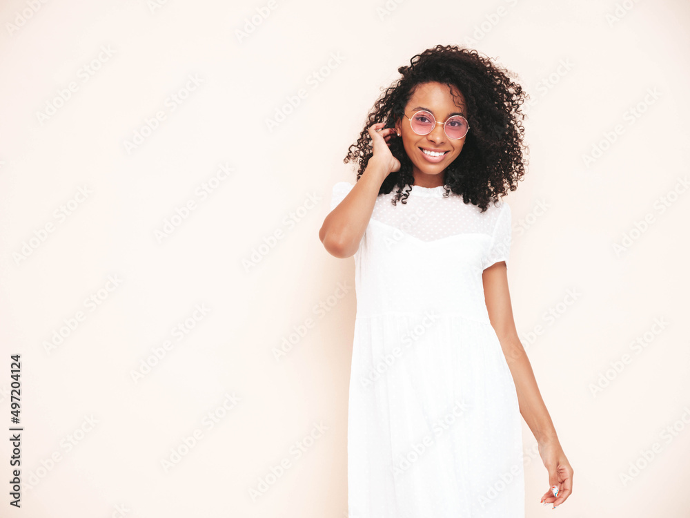 Beautiful black woman with afro curls hairstyle. Smiling model dressed in white summer dress. Sexy carefree female posing near wall in studio. Tanned and cheerful. Isolated Beautiful black woman with afro curls hairstyle. Smiling model dressed in white summer dress. Sexy carefree female posing near wall in studio. Tanned and cheerful. Isolated